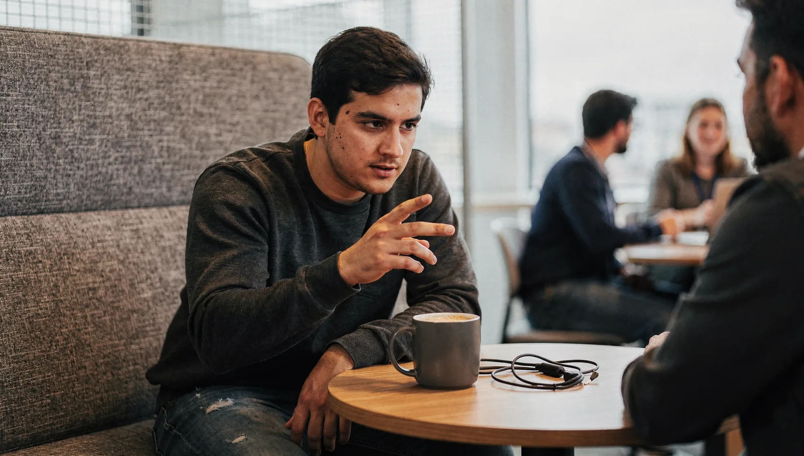 A man leans over an office table, gesturing with pinched fingers while speaking to an unseen colleague.