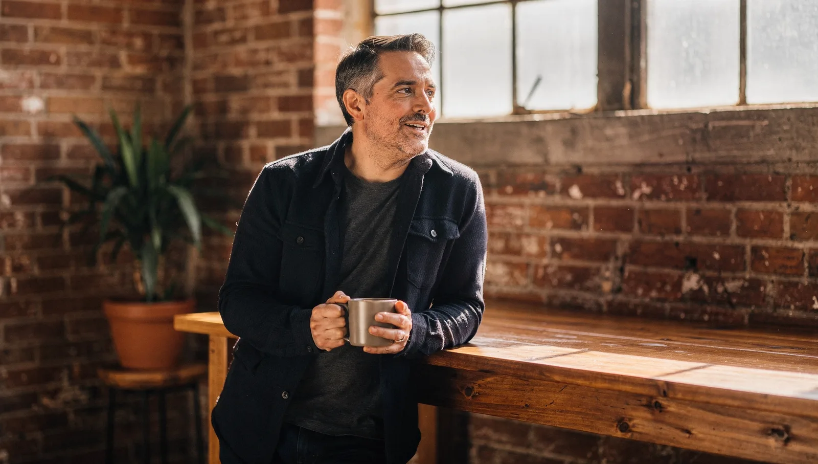 Man leaning against an oak workbench holding a coffee mug, listening in a sunlit industrial office nook.