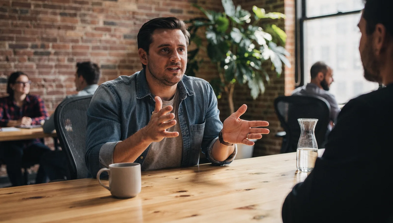 A person in a denim shirt gestures while speaking at a conference table in a sunlit loft office.