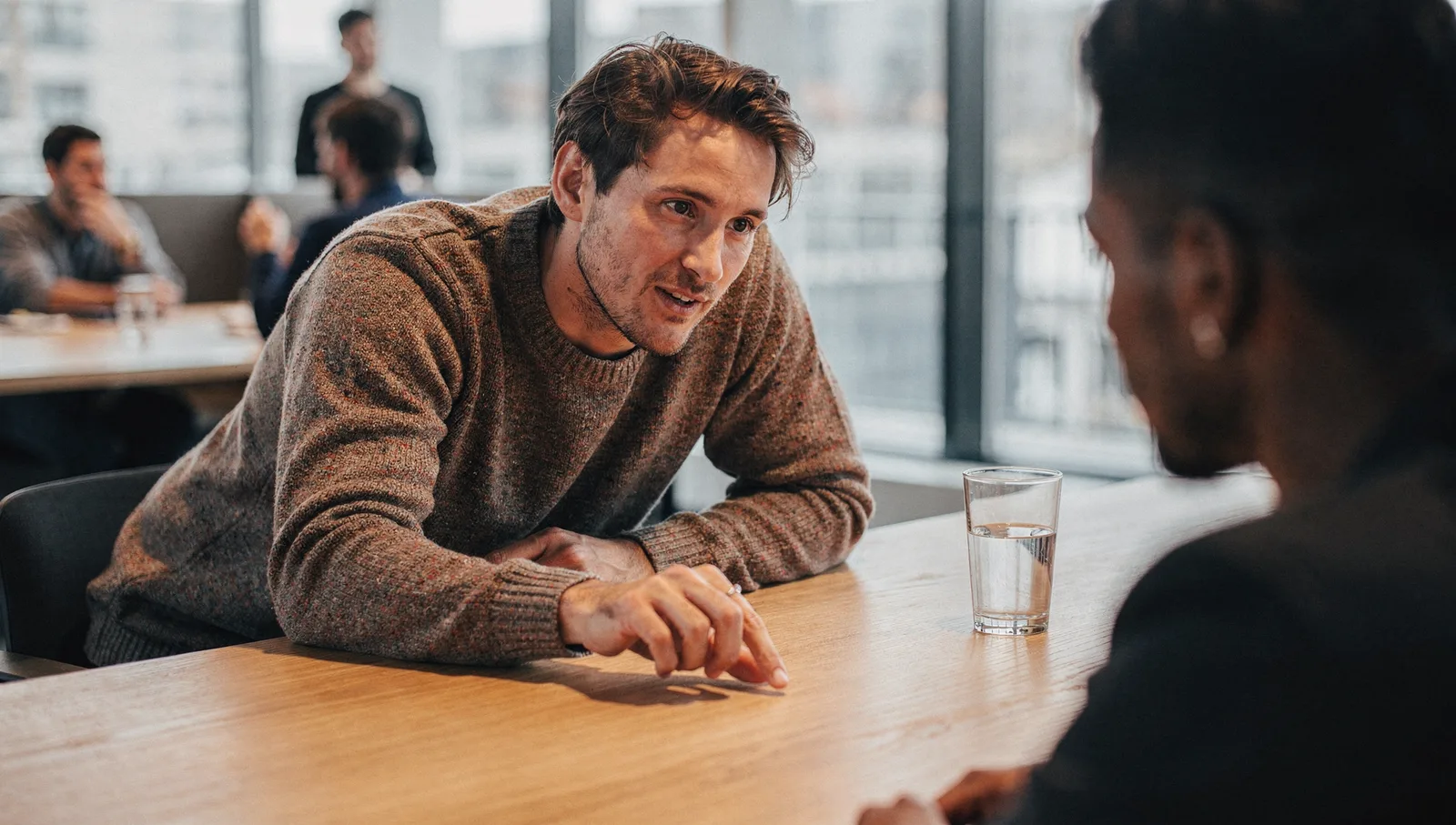 A man in a wool sweater leans across a wooden office desk, tapping the surface while speaking to a colleague.