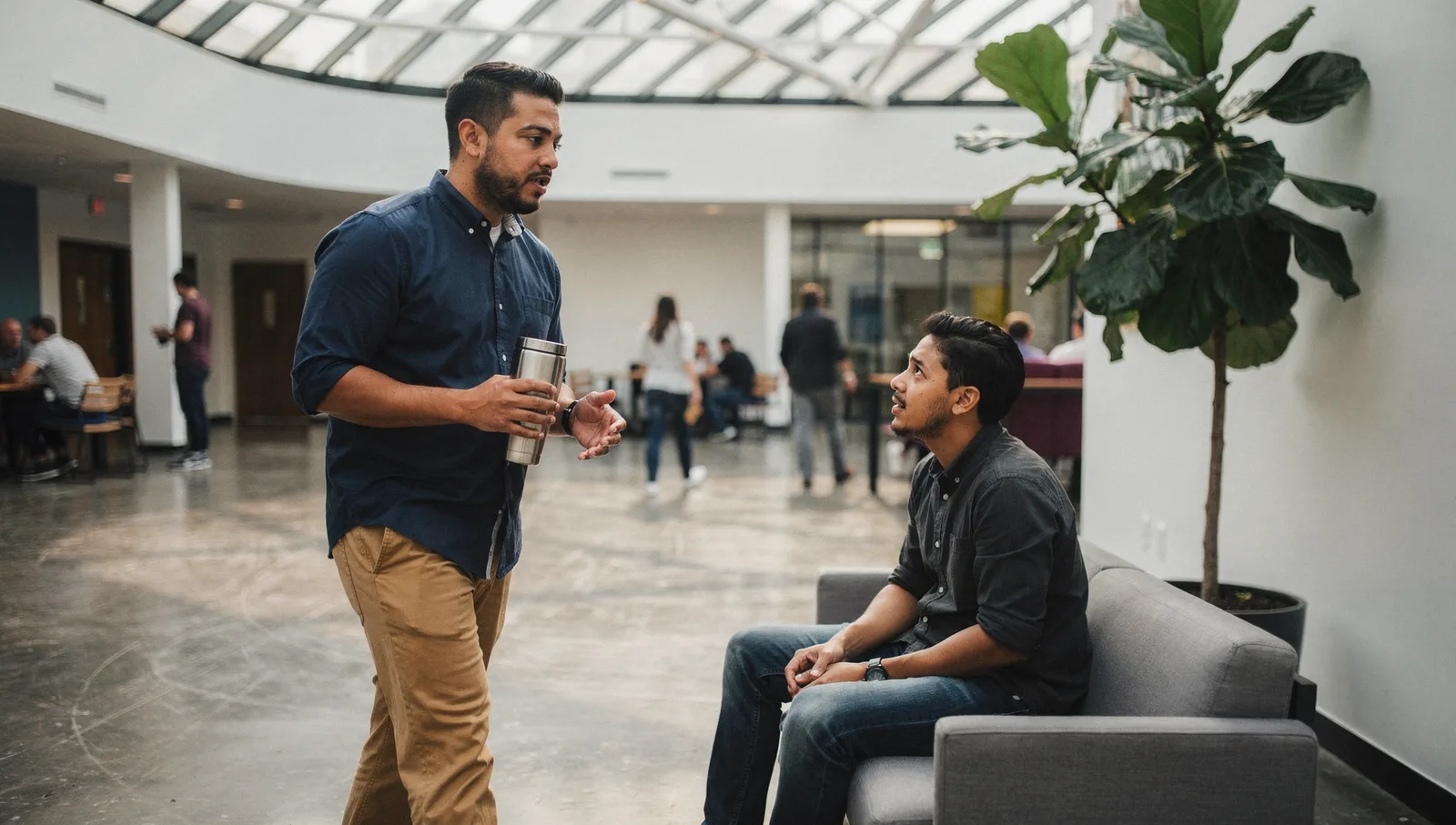 A man in a navy shirt holds a tumbler and walks past a seated colleague in a sunlit modern office atrium.