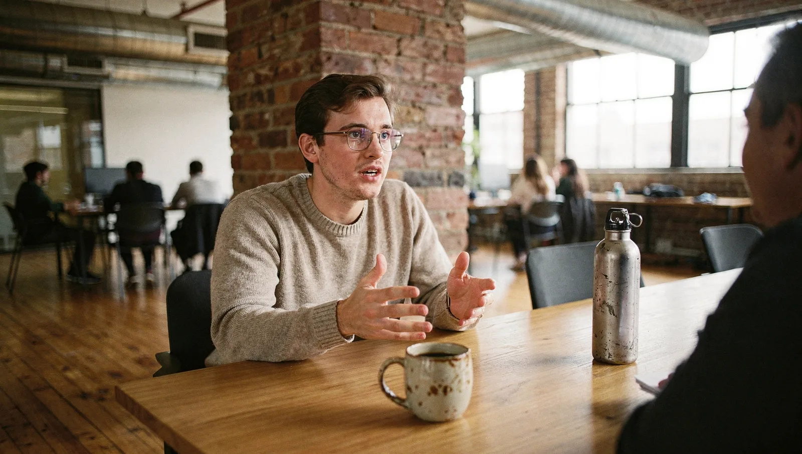 A person in spectacles and a knit sweater gestures emphatically across a wooden table in an open-plan office.