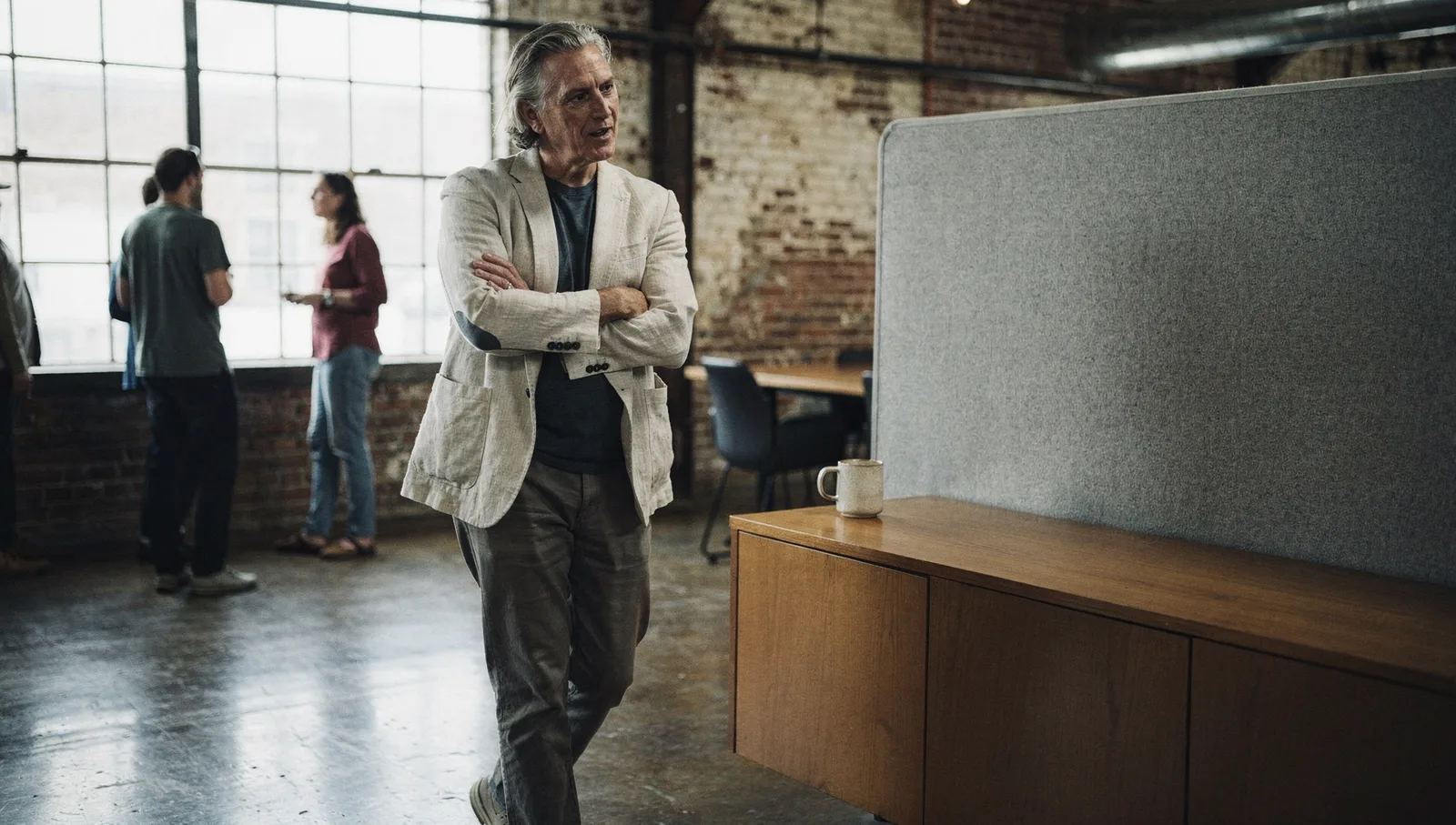 A professional in a linen blazer listening with crossed arms in an industrial studio meeting room in morning daylight.