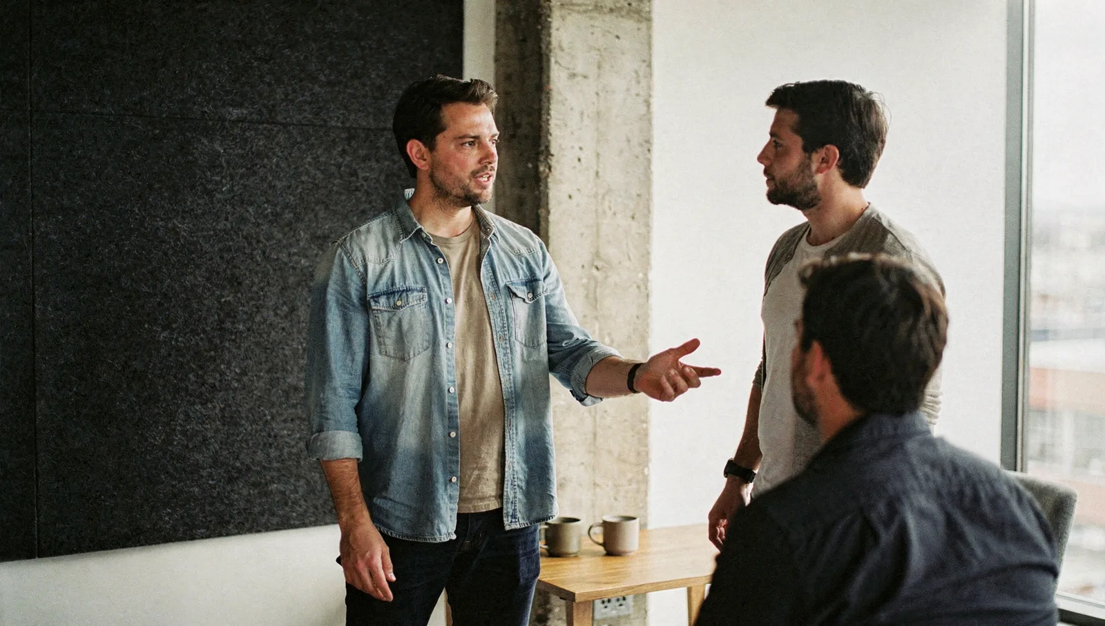 Two colleagues discuss strategy in a modern office breakout space with large windows and concrete pillars.